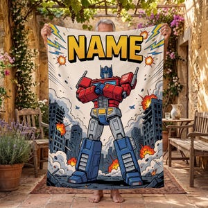 May include: A rectangular blanket featuring a comic book style illustration of a red and blue robot standing in a city with explosions. The word NAME is printed in large yellow letters at the top. The design includes blue lightning bolts.