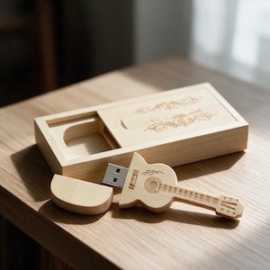 Puede incluir: Una memoria USB de madera clara con forma de guitarra acústica, que incluye una tapa extraíble y una caja rectangular de madera a juego con tapa deslizante y grabados florales decorativos en la superficie.