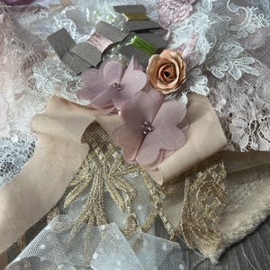 May include: A collection of craft supplies featuring white lace, beige fabric ribbons, and gold mesh netting. The arrangement includes two dusty pink fabric flowers with beaded centres, a small peach paper rose, and three spools of metallic thread.