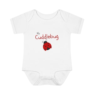 May include: A white short-sleeved baby bodysuit featuring the text My Cuddlebug in red and black lettering. Below the text is a graphic of a red ladybug with black spots and black antennae. The garment has a round neckline and leg openings.