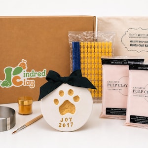 May include: A craft kit featuring a brown cardboard box with a Kindred Clay logo, two bags of creative pulp clay, a wooden rolling pin, a metal ring mold, a small gold paint jar, a paintbrush, a yellow letter stamp set, and a white clay paw print ornament.