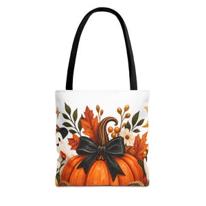 Peut inclure: Un sac fourre-tout blanc avec des anses noires orné d'une citrouille orange centrale agrémentée d'un grand nœud noir. Le motif comprend des feuilles d'automne, des petites fleurs blanches, des baies dorées et un petit fantôme avec un chapeau noir.