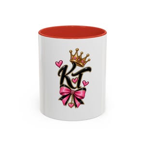 Peut inclure: Un mug en céramique blanche avec un intérieur et un bord rouges. L'avant présente les lettres KT en noir avec des contours dorés, surmontées d'une couronne dorée avec des gemmes roses. Un nœud rose et noir avec un petit cœur est en dessous.