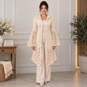 Puede incluir: Un cárdigan largo de ganchillo color crema con capucha, mangas largas acampanadas y diseño abierto en la parte delantera. La prenda presenta un patrón calado detallado y repetitivo en el cuerpo y las mangas, combinado con pantalones color crema.