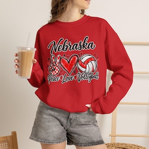 Op de afbeelding: Een rood sweatshirt met lange mouwen en een grafische print met de tekst Nebraska, Peace, Love, Volleyball. Het ontwerp bevat een rood-wit geblokt vredesteken, een rood hart en een volleybal, geaccentueerd met kleine rode sterretjes en hartjes.