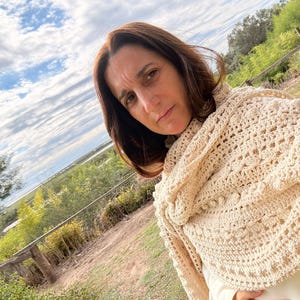 May include: A cream-colored crochet shawl featuring an intricate pattern of openwork stitches, textured bobbles, and a decorative border. The shawl is draped over the shoulders, showcasing the detailed yarn work and the soft, neutral tone of the fabric.