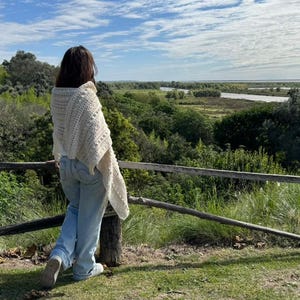 Puede incluir: Un chal de ganchillo texturizado de color crema drapeado sobre los hombros, combinado con pantalones vaqueros de mezclilla azul claro. El chal presenta un patrón repetitivo de puntos y una apariencia de tejido suave, sobre una camiseta blanca.
