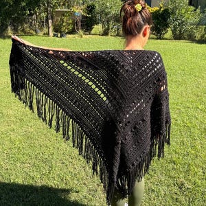 May include: A black triangular crochet shawl featuring a textured pattern with bobble stitches, openwork sections, and long fringe trim along the edges. The shawl is draped over the shoulders, showcasing the intricate yarn work and dark color.