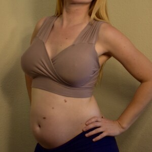 May include: A beige wrap-style nursing bra with wide straps. The bra is paired with navy blue bottoms. The woman is standing with her hand on her hip.