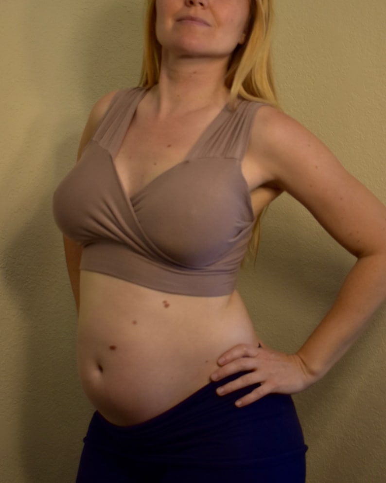 May include: A beige wrap-style nursing bra with wide straps. The bra is paired with navy blue bottoms. The woman is standing with her hand on her hip.