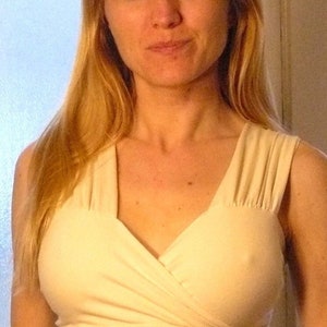 May include: A woman with long blonde hair is wearing a cream-colored, sleeveless top with a V-neck and a wrap-style design.