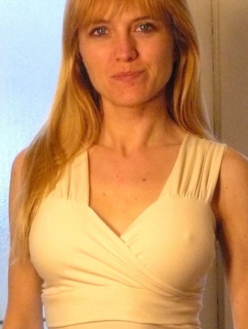 May include: A woman with long blonde hair is wearing a cream-colored, sleeveless top with a V-neck and a wrap-style design.