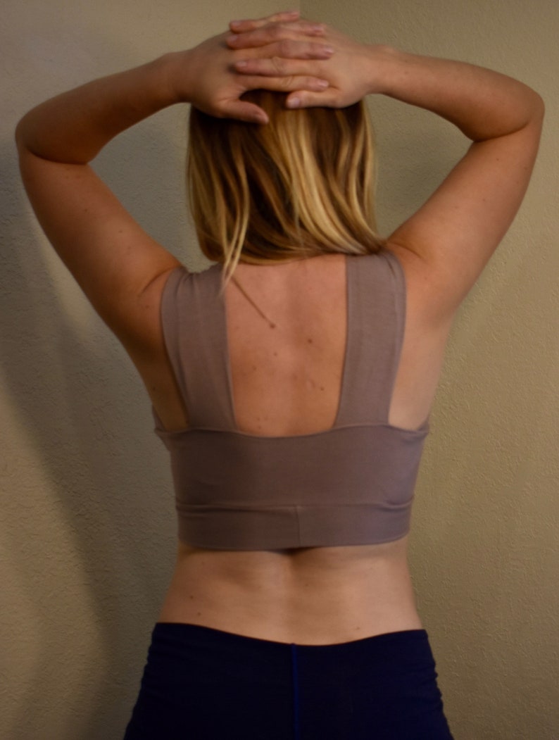 May include: A taupe sports bra with wide straps and a scoop neckline. The bra is paired with navy blue athletic shorts. The person's arms are raised above their head, with their hands clasped together.