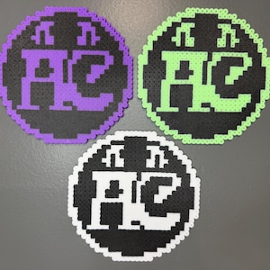 May include: Three circular pixel art coasters made of fused plastic beads. Each coaster features a black background with the letters AC and two musical notes above them. One coaster has a purple border and design, one is lime green, and one is white.