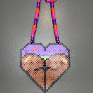 Puede incluir: Un colgante en forma de corazón hecho de cuentas de plástico fusionadas, con una sección superior morada con una pequeña cruz rosa y una sección inferior marrón con una línea vertical negra. El collar está hecho de cuentas moradas y naranjas alternadas.
