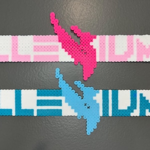 May include: Two rectangular perler bead creations featuring the text ILLENIUM in a block font. The top piece is white with pink lettering and a pink phoenix emblem. The bottom piece is white with blue lettering and a blue phoenix emblem.