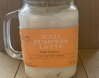 Könnte beinhalten: Eine Kerze im durchsichtigen Einmachglas mit Henkel und silbernem Metalldeckel. Die Kerze enthält weißes Sojawachs und hat ein orangefarbenes Etikett mit dem Text ICED PUMPKIN LATTE, funkE Creations, 100% SOY WAX CANDLE und 13 OZ.