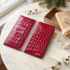 May include: A deep red, rectangular wallet with a glossy crocodile-embossed texture. The interior features multiple card slots and a central compartment, with the word Maktub embossed in gold lettering on the lower right corner.