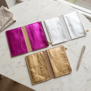 May include: Three rectangular passport holders in metallic fuchsia, silver, and gold, each featuring interior card slots and a small gold-tone zipper pull. A beige pen lies next to the gold holder. Each holder has a small embossed name.