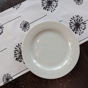 May include: A white ceramic dinner plate with a slightly raised rim sits on a white table runner with a black dandelion pattern. The table runner is on a dark wood surface.