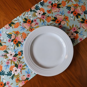 May include: A white ceramic dinner plate sits on a blue and orange floral table runner. The runner has a light blue background with a repeating pattern of flowers in shades of orange, pink, and white.