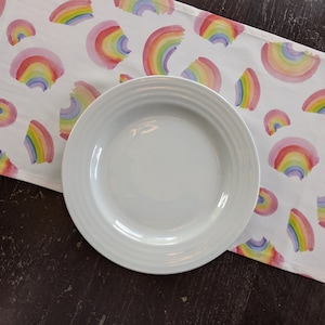 May include: A white ceramic plate rests on a white table runner decorated with watercolor rainbows in shades of pink, yellow, blue, and purple. The runner is on a dark brown surface. The plate has a diameter of approximately 10 inches.