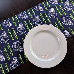 May include: A navy blue table runner featuring a repeating pattern of Seattle Seahawks football helmets and text. A white plate sits on the runner. The runner is made of fabric.