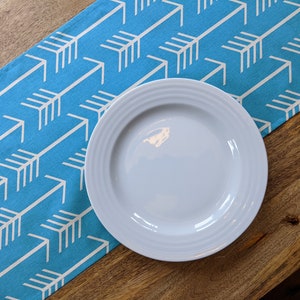May include: A white ceramic dinner plate with a slightly raised rim sits on a blue and white table runner with a repeating arrow pattern.