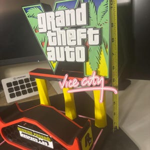 May include: A 3D-printed Grand Theft Auto Vice City sign featuring a large V, palm trees, and pink text. A yellow measuring tape shows the sign is approximately 11 inches tall. A black and orange base displays the text Mission Passed Respect +99.