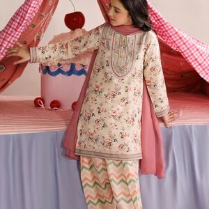 May include: A long-sleeved tunic featuring a cream base with a pink and green floral print, paired with matching trousers displaying a pink and green chevron pattern. The outfit includes a solid pink scarf draped over the shoulders and shoulders.
