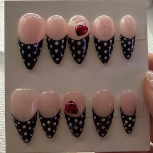 May include: A set of ten press-on nails arranged on a white display card. Each nail features a light pink base with a black tip decorated with white polka dots. Two of the nails feature a small, three-dimensional red ladybug accent on the tip.