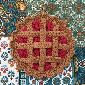 May include: A round, crocheted potholder designed to resemble a cherry pie. It features a deep red, textured center representing the fruit filling, overlaid with a tan, lattice-style crust pattern and a scalloped edge with a small hanging loop.