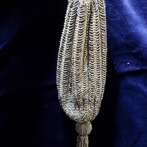 May include: A drawstring pouch bag covered in vertical rows of metallic silver and dark grey beads. The bag features a gathered top with a matching cord closure and a beaded spherical accent at the base, which transitions into a long, dangling beaded tassel.