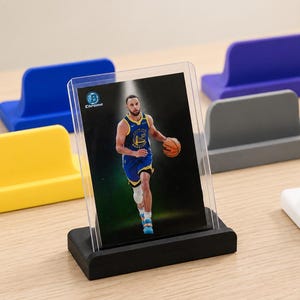 May include: A collection of L-shaped plastic display stands in various colors including black, white, yellow, blue, purple, and gray. A clear plastic card holder containing a basketball trading card is positioned in the black display stand.