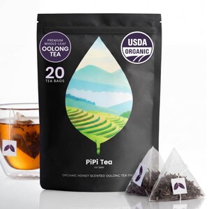 May include: A black pouch of PiPi Tea containing 20 organic honey scented oolong tea bags. The packaging features a leaf-shaped illustration of green terraced hills and mountains. A glass cup of brewed tea and two pyramid tea bags are visible.