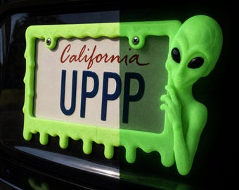 Peut inclure: Un cadre de plaque d'immatriculation vert néon vif avec un design liquide dégoulinant et une figurine d'extraterrestre en trois dimensions sur le côté droit. Le cadre entoure une plaque californienne avec le texte UPPP en bleu foncé.