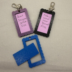 May include: Three rectangular scalloped frames with metal lobster clasps. The purple frame contains a pink insert with the text Chart, Designer, Stitch, Size. The black frame has a pink insert with the text Fabric, count, color, Brand, Floss. A blue frame is empty.