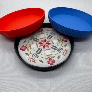 May include: A set of three round, ribbed plastic trays in red, blue, and black. The black tray contains a circular piece of white fabric featuring a cross-stitch pattern of red flowers, green leaves, and grey scissors arranged in a symmetrical design.