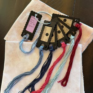 May include: A set of embroidery floss samples on black scalloped plastic tags held by a metal ring. The tags feature labels with text including Weeks Dye Works and color names. The floss colors are light blue, dark blue, pink, and deep red.