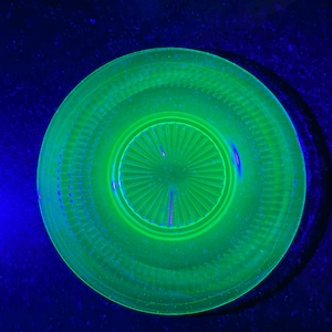 May include: A circular glass plate glowing with a bright neon green fluorescence under ultraviolet light. The plate features a central sunburst pattern of radiating lines surrounded by a textured rim with a series of small, evenly spaced indentations.