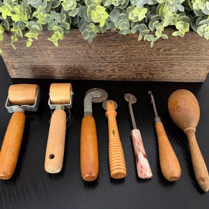 May include: A collection of six vintage crafting tools including two wooden rollers with metal frames, a metal tracing wheel with a wooden handle, a small serrated wheel, a metal hook tool with a wooden handle, and a wooden darning egg.