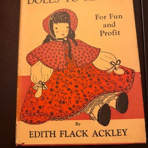 May include: A hardcover book titled Dolls to Make For Fun and Profit by Edith Flack Ackley. The cover features a graphic illustration of a doll wearing a red bonnet and a red dress with a black floral pattern, set against a light orange background.