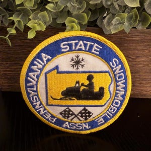 May include: A circular embroidered patch with a yellow border and a blue outer ring featuring the white text Pennsylvania State Snowmobile Assn. The center shows a yellow map of Pennsylvania with a black snowmobile, a snowflake, and two flags.