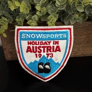 Puede incluir: Un parche bordado en forma de escudo con fondo blanco y bordes rojos y azules. Presenta el texto SNOWSPORTS, HOLIDAY IN AUSTRIA y 19 73 en hilo rojo y azul. Debajo hay un gráfico de una montaña azul con dos pájaros en blanco y negro.
