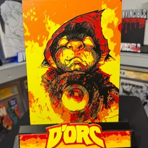 May include: A rectangular print featuring a stylized illustration of a hooded figure with glowing yellow eyes against a vibrant orange and yellow background. Below the print, a black stand displays the text D'ORS in bold yellow letters.