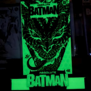 Op de afbeelding: Een oplichtend groen en zwart displaybord met de tekst Absolute Batman aan de boven- en onderkant. De centrale afbeelding toont een gestileerd, scherpgetand wezengezicht met een lange tong, verlicht in fel neongroen tegen een donkere achtergrond.