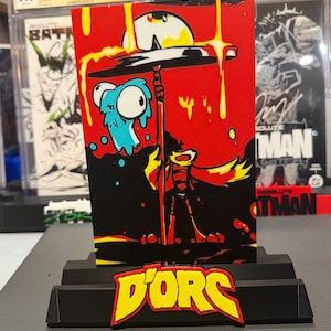May include: A rectangular art print featuring a stylized character holding a staff under a dripping yellow sun against a red background, with a blue creature nearby. The print is displayed on a black stand with a yellow and red D'ORC logo attached.