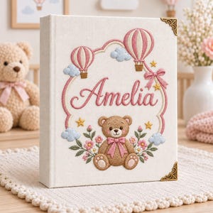 May include: A cream-coloured fabric-covered photo album featuring intricate embroidery of a brown teddy bear with a pink bow, two pink and white hot air balloons, light blue clouds, yellow stars, and floral accents, with the name Amelia in pink script.