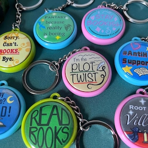 May include: A collection of eight circular keychains with metal rings, each featuring unique colorful designs and text including "Sorry. Can't. BOOKS. Bye.", "FANTASY because reality is boring", "STAY BOOKISH", "I'm the PLOT TWIST", "#AntiHeroSupporter", "Why Sleep? READ!", "READ BOOKS", and "ROOT FOR Villians".