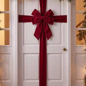 May include: A white door decorated with a large, deep red velvet ribbon and bow, arranged in a cross pattern to resemble a gift wrap. The ribbon extends vertically and horizontally across the door panels, centred with a full, multi-layered bow.